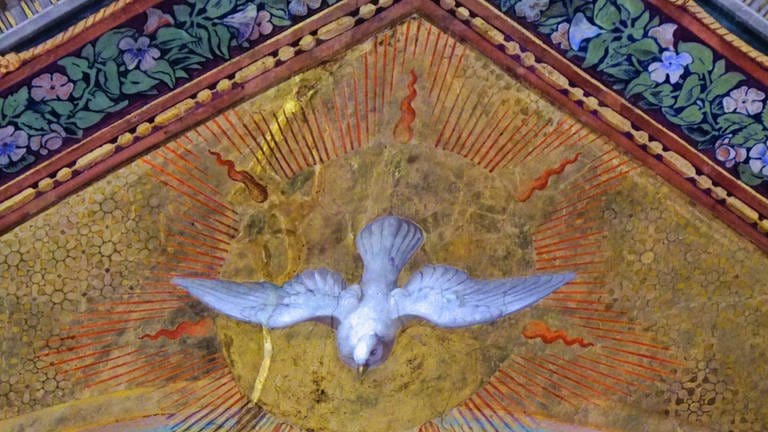 Eine weiße Taube vor einem gemalten Himmel einer Kirchendecke.