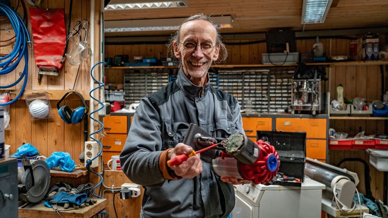 Elektriker Heinrich Jung streckt ein defektes Elektrogerät lächelnd in die Kamera.