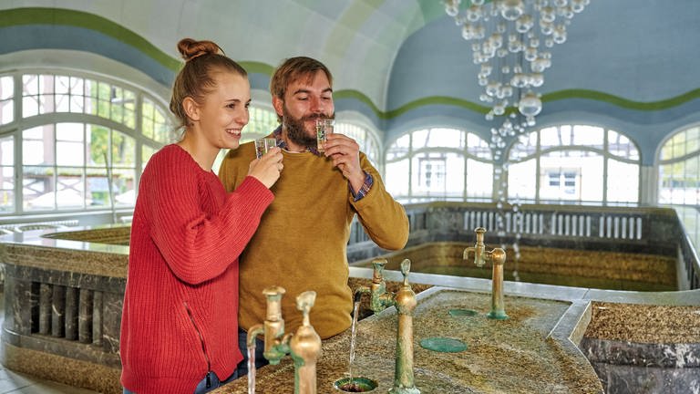 Ein Paar trinkt Solewasser aus den Hähnen in der Brunnenhalle in Bad Münster am Stein