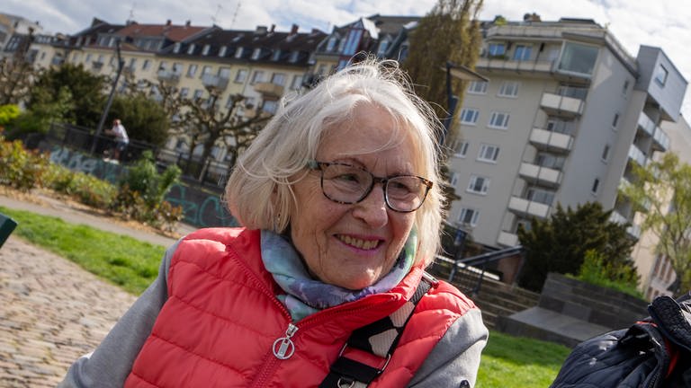 Eine ältere Dame mit Brille und pinker Jacke sitzt auf einer Bank und lächelt zur Seite.