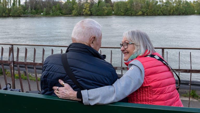 Eine ältere Dame legt einem älteren Herrn den Arm um die Schulter und lächelt ihn an.
