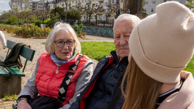 Drei Personen sitzen auf einer Bank. Von einer älteren Dame und einem älteren Herrn kann man das Gesicht sehen. Sie schauen auf die dritte Person, die man nur von hinten sieht mit rosa Mütze und Jacke. 
