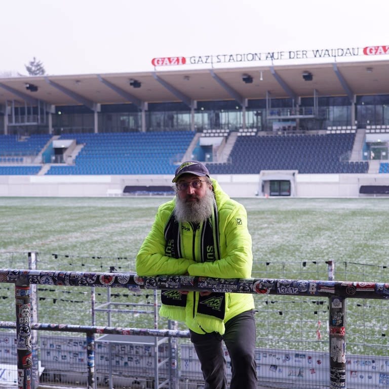 David trägt Football-Fanartikel der Stuttgart Surge. Er ist im GAZi-Stadion (Stuttgart Surge Stadion) und steht im B-Block. Er lehnt sich über ein Geländer und schaut in die Kamera. Im Hintergrund ist das Stadion mit der Haupttribüne zu sehen.