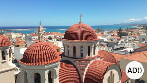 Im Vordergrund die Kirche Tessaron Martyron in der Hafenstadt Rethymno im Norden der griechischen Insel Kreta, dahinter in leuchtendem blau das Mittelmeer, rechts unten das Icon AD, das kennzeichnet, dass ein Film eine Audiodekription hat