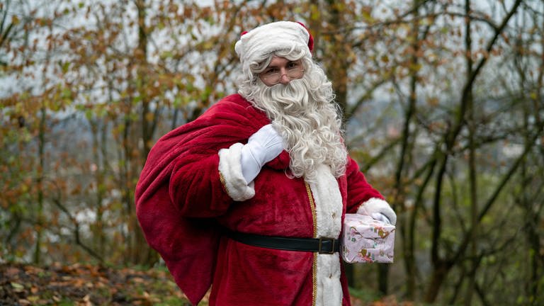 Mann in Nikolauskostüm mit weißem Bart und Geschenk vor Bäumen.