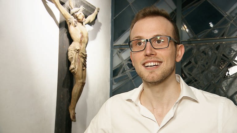 Junger Mann mit Brille steht in einer Kirche vor einem Holzkreuz. (Foto: SWR)