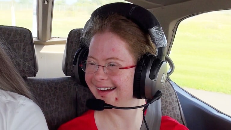 Junge Frau sitzt im Flugzeug, trägt Kopfhörer und lächelt. 