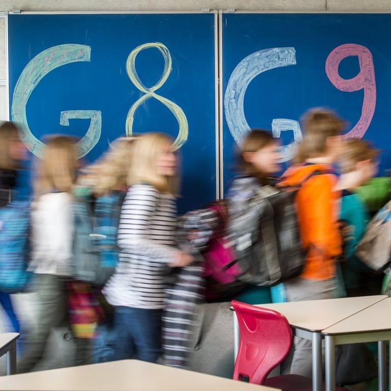 An einer Tafel in einem Klassenzimmer steht links "G8" und rechts "G9" geschrieben. Schülerinnen und Schüler laufen vor der Tafel auf die rechte Seite der Tafel, auf der "G9" steht. 