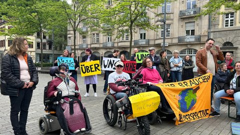 Menschen mit Rollstühlen und ohne haben sich in Heidelberg versammelt. Sie halten Banner und protestieren für mehr Inklusion.
