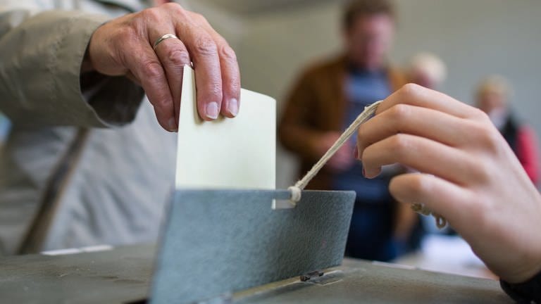Hand wirft einen gelben Wahlzettel in eine Wahlurne. Am 14. März 2021 ist Landtagswahl in Rheinland-Pfalz 