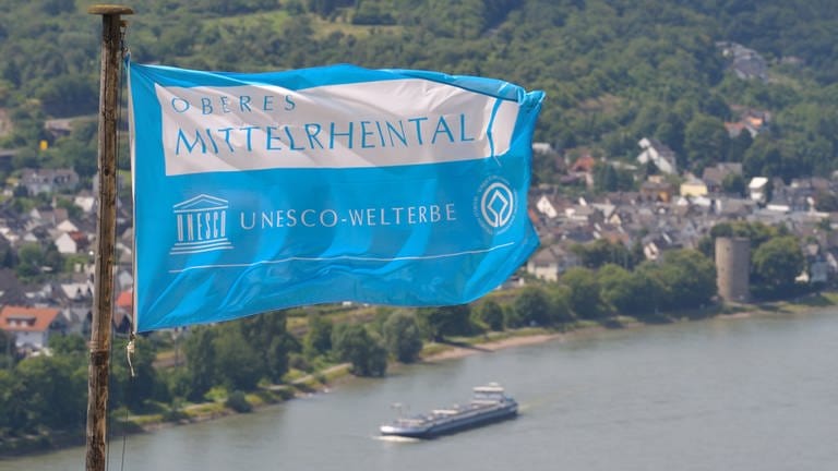 Eine Fahne mit der Aufschrift "Oberes Mittelrheintal - UNESCO-Welterbe" weht am 27.07.2009 über der Marksburg in Braubach bei Koblenz, während im Hintergrund ein Frachtschiff auf dem Rhein zu sehen ist.
