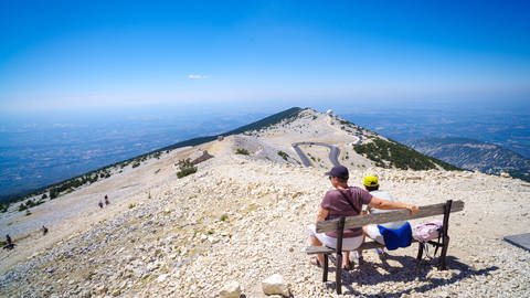 Zwei Tourist*innen auf einer Band auf dem Mont Ventoux mit Blick über die umliegende Landschaft