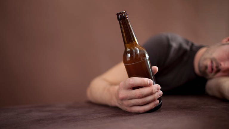 Ein Mann liegt mit dem Oberkörper an der Tischkante und hält eine Flasche Bier in der Hand