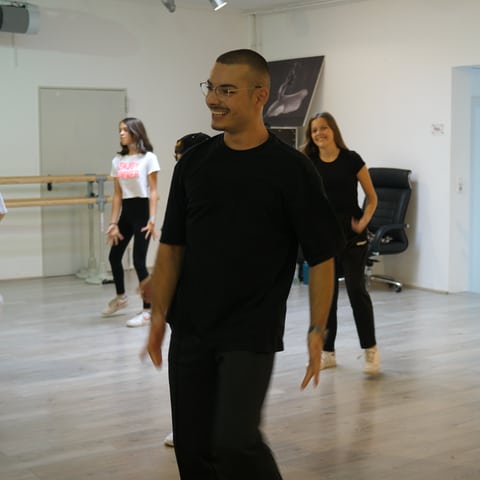 Tanzlehrer Deniz ist in einem Tanzstudio. Dort bringt er Kindern das Tanzen bei. Deniz macht eine Tanzübung vor und lacht dabei. Im Hintergrund sind Kinder zu sehen, die Deniz’ Tanzbewegung nachmachen. 