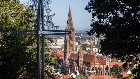 Ein stilisiertes Kreuz steht auf dem Freiburger Schlossberg während im Hintergrund das Münster zu sehen ist.