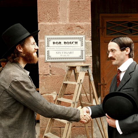 Zwei dunkel gekleidete Männer geben sich die Hand. Beide tragen Hut und Brille. Hinter ihnen sieht man mittig an einer Mauer ein Schild auf dem steht: "Rob Bosch, Werkstätte für Feinmechanik & Elektrotechnik, Stuttgart." (Foto: SWR, Peter Trinks)
