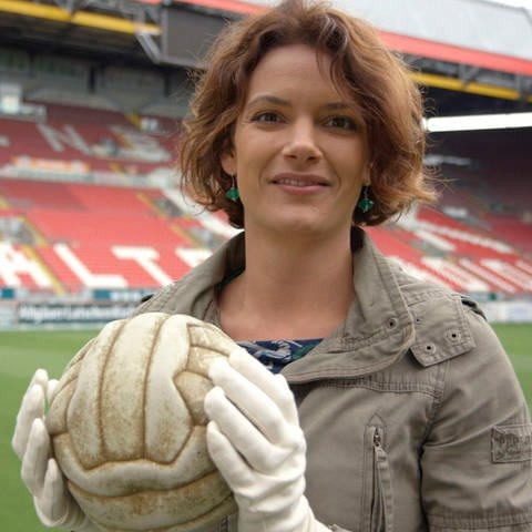Lena Ganschow steht in einem leeren Stadion. Sie trägt Handschuhe und hält einen alten Fußball in den Händen. (Foto: SWR, Roland Breitschuh)
