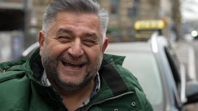 Iraklis ist Taxifahrer in Stuttgart. Er steht in der Stadt und lächelt herzlich in die Kamera.  (Foto: SWR)