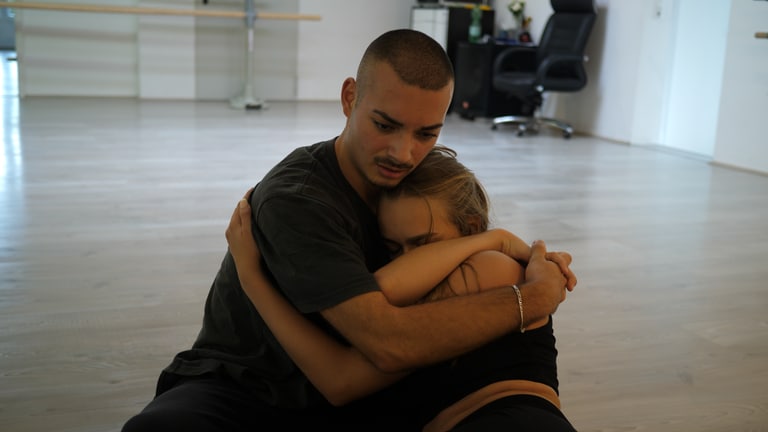 Tanzlehrer Deniz tanzt zusammen mit einer Tanzpartnerin. Die beiden sitzen auf dem Boden und halten sich in den Armen. Deniz’ Blick ist gefühlvoll und emotional.