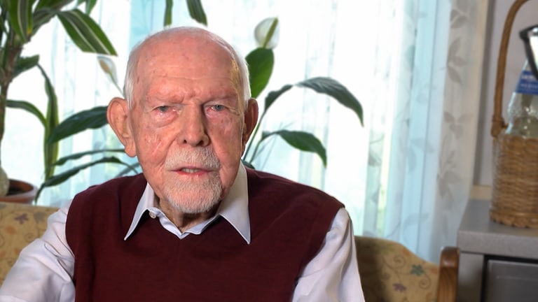Helmut ist 95 Jahre alt, sitzt auf seinem Sofa und blickt in die Kamera.