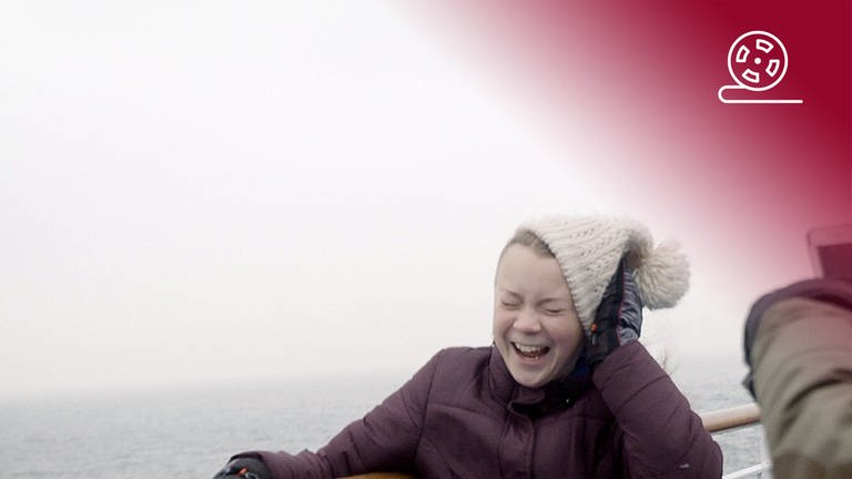 Foto zum Dokumentarfilm Ich bin Greta: Während der Segelreise zum Klimagipfel hält Greta sich bei einem Sturm lachend ihre Mütze fest und wird dabei mit einem Smartphone aufgenommen, rotes Overlay mit Icon Filmrolle