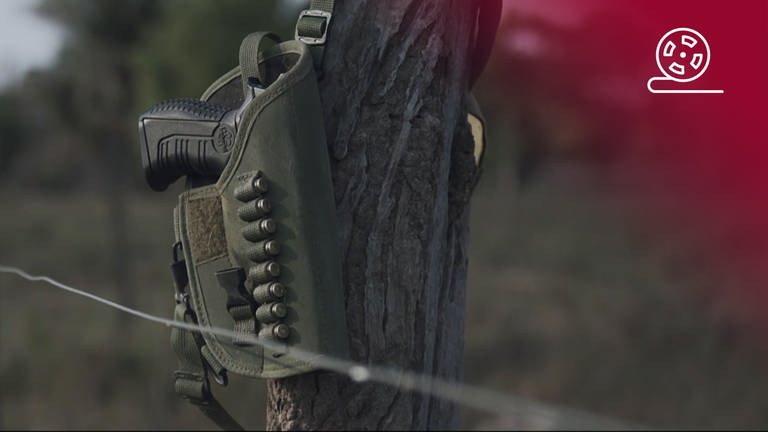 Standbild aus dem Dokumentarfilm Blutige Kohle: Waffenholster hängt an einem Zaunpfahl, rotes Overlay mit Icon Filmrolle