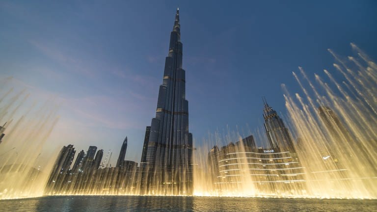 Burj Khalifa hebt sich dunkel vom blauen Abendhimmel ab. Im Vordergrund schießen golden beleuchtete Wasserfontänen in die Höhe.