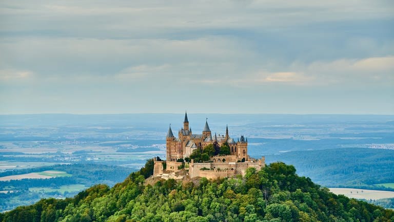 Die Burg Hohenzollern liegt auf der Schwäbischen Alb in Baden-Württemberg. Rundum eine grüne Landschaft und die Stadt mit blauen Himmel im Hintergrund.