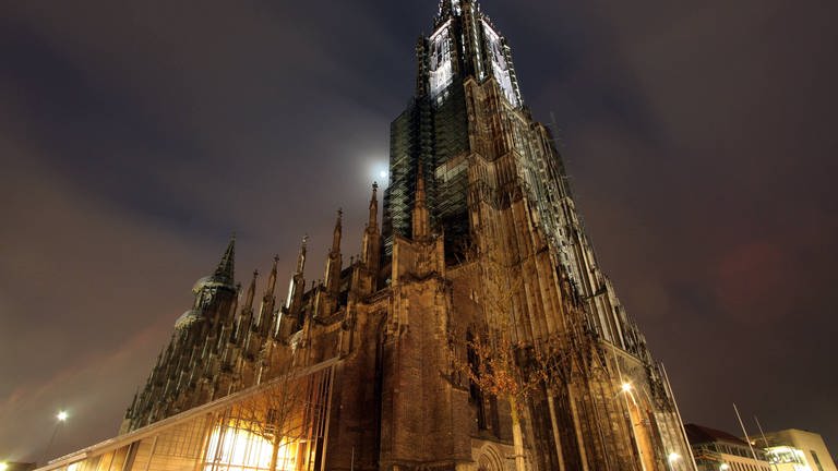 Das Ulmer Münster hebt sich durch eine gelbe Beleuchtung vom blau-schwarzen Abendhimmel ab. Es hat den höchsten Kirchturm der Welt.