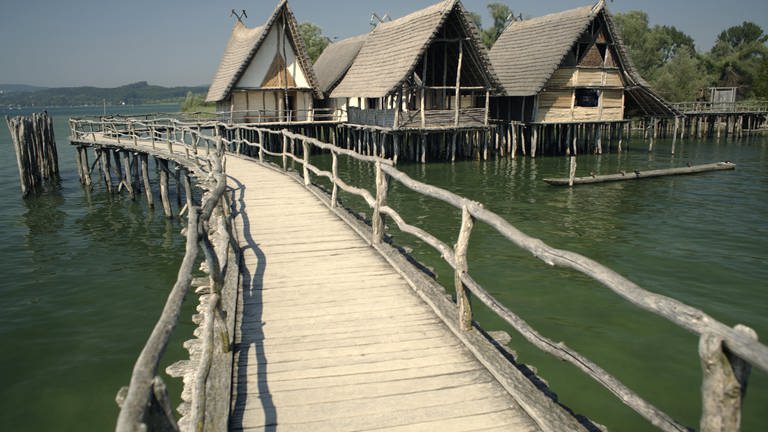 Die Pfahlbauten in Unteruhldingen sind Häuser aus Holz auf Stelzen, die auf dem Wasser gebaut sind. In der Mitte ist ein Holzsteg.