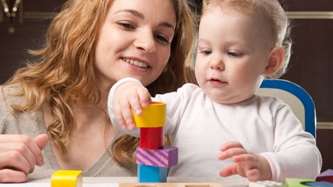 Ein Baby baut einen Turm, die Mutter beobachtet es. In der Phase der Ein-Wort-Äußerungen baut das Kind seinen Wortschatz aus. Ab etwa zwei Jahren können Kinder Wörter kombinieren und erste Mehrwort-Äußerungen produzieren: Turm bauen.  (Foto: Colourbox)