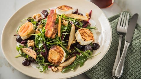 Ziegenkäsetaler auf Salat mit etwas Speck und Trauben, daneben Besteck und 