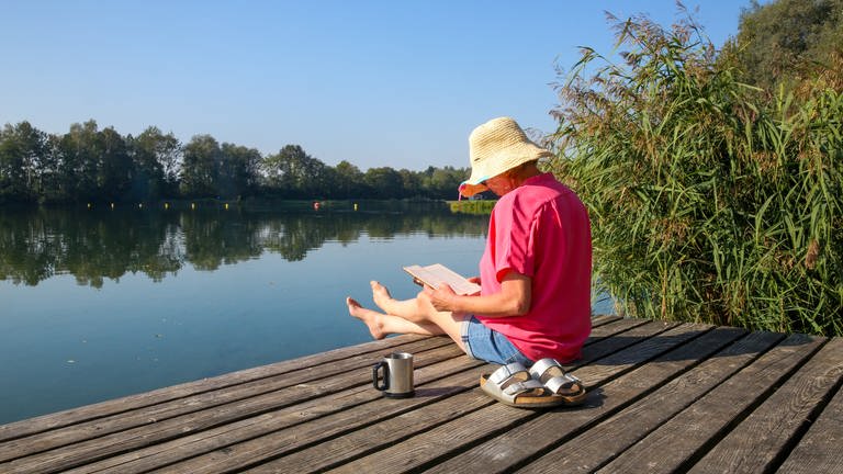 Eine Frau sitzt mit einem Buch in der Hand und einer Tasse Kaffe auf einer Badeplattform in der Sonne.