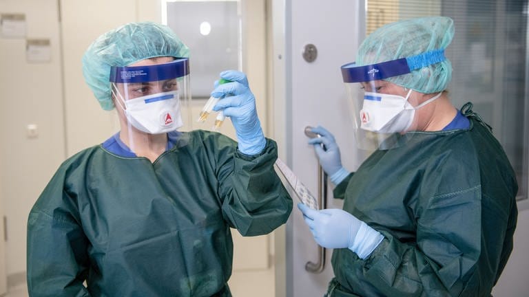 Zwei Krankenpflegerinnen betrachten vor einem Krankenzimmer in Schutzkleidung und hinter einer Atemmaske zwei Abstrichröhrchen und den dazu gehörenden Virologieschein. 