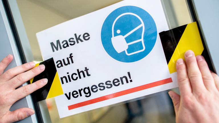 Eine Person klebt ein Schild mit der Aufschrift "Maske auf nicht vergessen!" an die Glasscheibe einer Zwischentür.