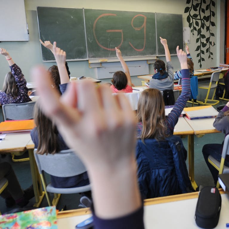 Schülerinnen und Schüler sitzen in einem Unterrichtsraum und heben ihre Finger, an der Tafel steht "G9". 