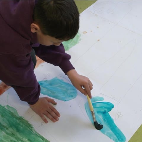 Ein Junge malt mit Wasserfarben auf dem Boden ein Selbstporträt aus. In der LEA in Ellwangen malen geflüchtete Kinder. Es ist zugleich Therapie und Beschäftigung.