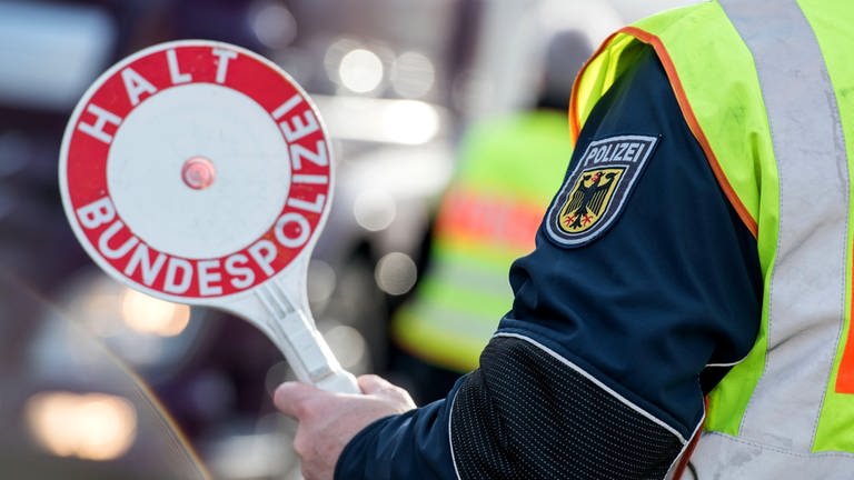 Ein Polizist mit gelber Weste hält eine rot-weiße Kelle mit der Aufschrift "Halt, Bundespolizei" hoch.