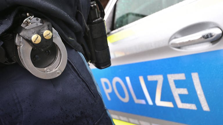 Ein Polizist mit Handschellen und einer Pistole am Gürtel steht vor einem Streifenwagen