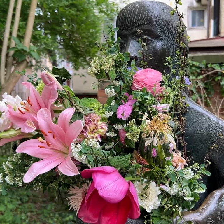 Man sieht eine Skulptur in einer Gasse in Tübingen. Sie hat einen großen Blumenstrauß in den Armen. Man nennt sie die "Sitzende". In Tübingen wundert man sich: Wer kümmert sich so liebevoll um die "Sitzende" und schenkt ihr Blumen? 