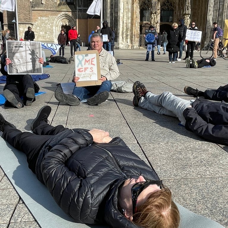 Menschen liegen auf dem Münsterplatz in Ulm, im Hintergrund das Münster. Sie demonstrieren im Liegen, um auf die Krankheit MECFS aufmerksam zu machen. Betroffene sind oft zu erschöpft, um das Haus zu verlassen.