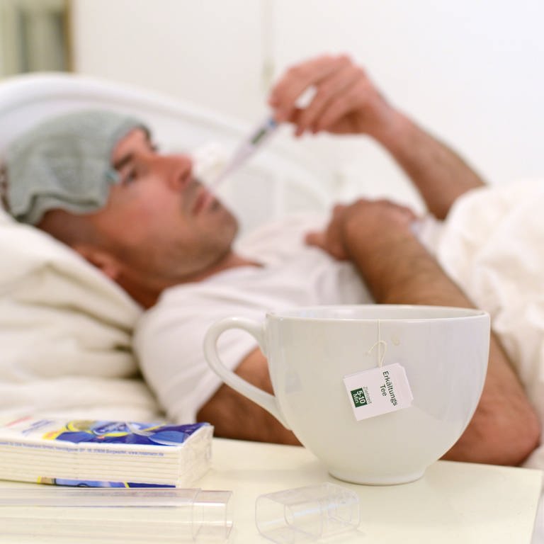 Ein kranker Mann mit Waschlappen zur Kühlung auf der Stirn liegt im Bett und misst Fieber - im Vordergrund steht eine Teetasse.