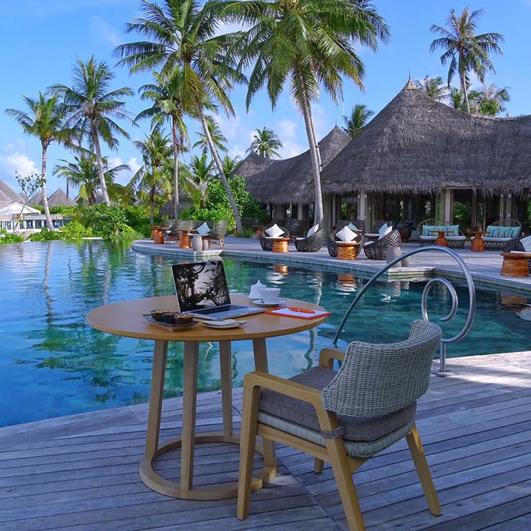 Ein Arbeitsplatz mit Laptop steht auf einem Café-Tisch mit Sessel neben einem Pool, umringt mit Palmen und Hütten einer Hotel-Anlage auf den Malediven. (Foto: dpa Bildfunk, picture alliance/dpa/The Nautilus Maldives | -)