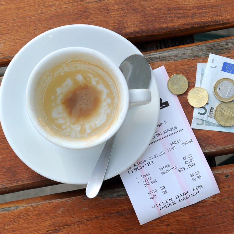 Münzen, ein Geldschein und eine Rechnung liegen neben einer leeren Kaffeetasse auf einem Tisch in einem Restaurant.