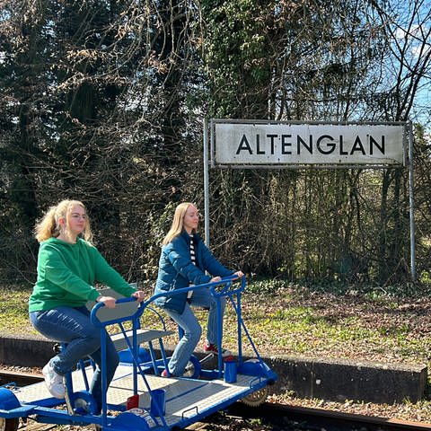 Zwei junge Frauen fahren auf einer Draisine durch die Landschaft, hinter ihnen das Ortsschild von Altenglan (Foto: SWR)