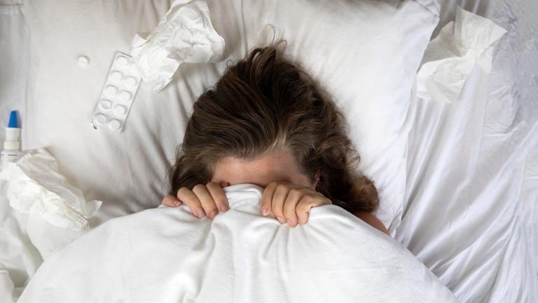 Grippekranke Frau hat sich im Bett die Decke über den Kopf gezogen, daneben Tablettenblister.