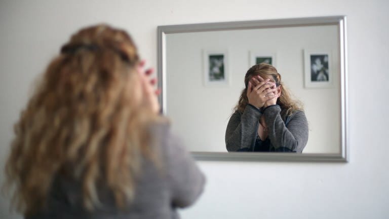 Eine Frau steht vorm Spiegel und hält sich verzweifelt die Hände vors Gesicht, sie ist nicht zu erkennen.