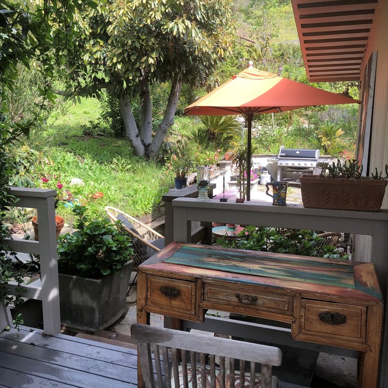 Kleiner Holz-Schreibtisch auf der Terrasse, Blick in den Garten mit alten Bäumen, Blumen und auf einen orange-farbenen Sonnernschirm (Foto: SWR, privat)