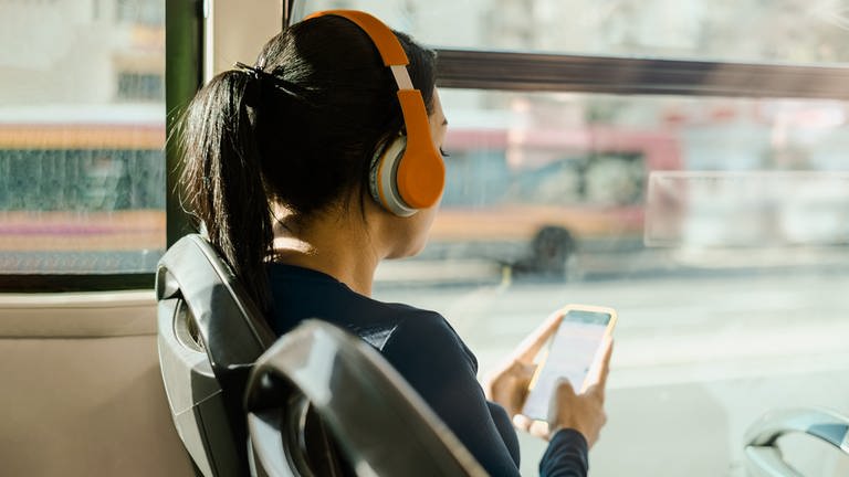 Eine Frau sitzt im Bus und hört mit Kopfhörern etwas über ihr Handy 