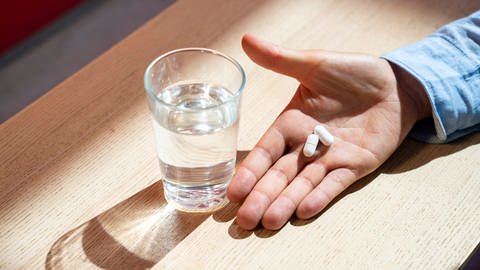 2 Tabletten auf einer Handfläche; daneben ein Wasserglas: Sterbewillige können einen Angehörigen, Bekannten oder eine Ärztin bitten, eine tödliche Substanz zu beschaffen. Das Gift muss der Sterbewillige selbst nehmen (Symbolfoto)